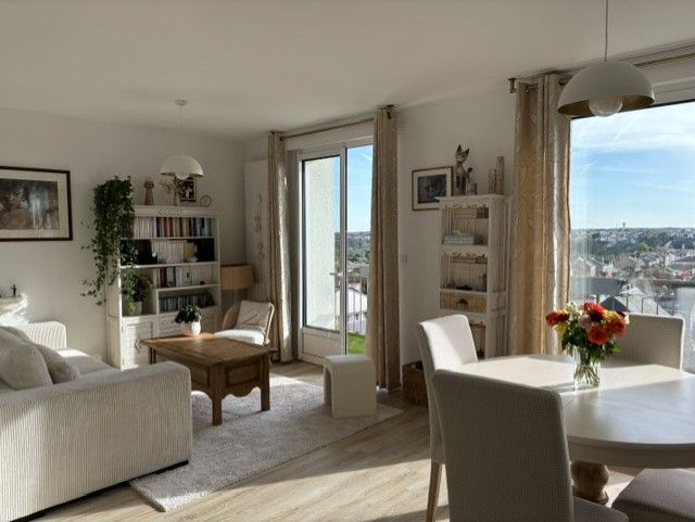 Salon-salle à manger lumineux avec meubles blancs, portes-fenêtres ouvertes sur le balcon et vase de fleurs sur la table