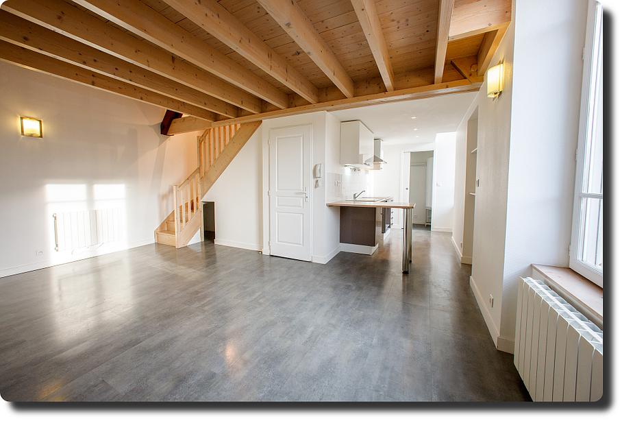 Intérieur de loft vide avec poutres apparentes, sol gris, murs blancs et une grande fenêtre.