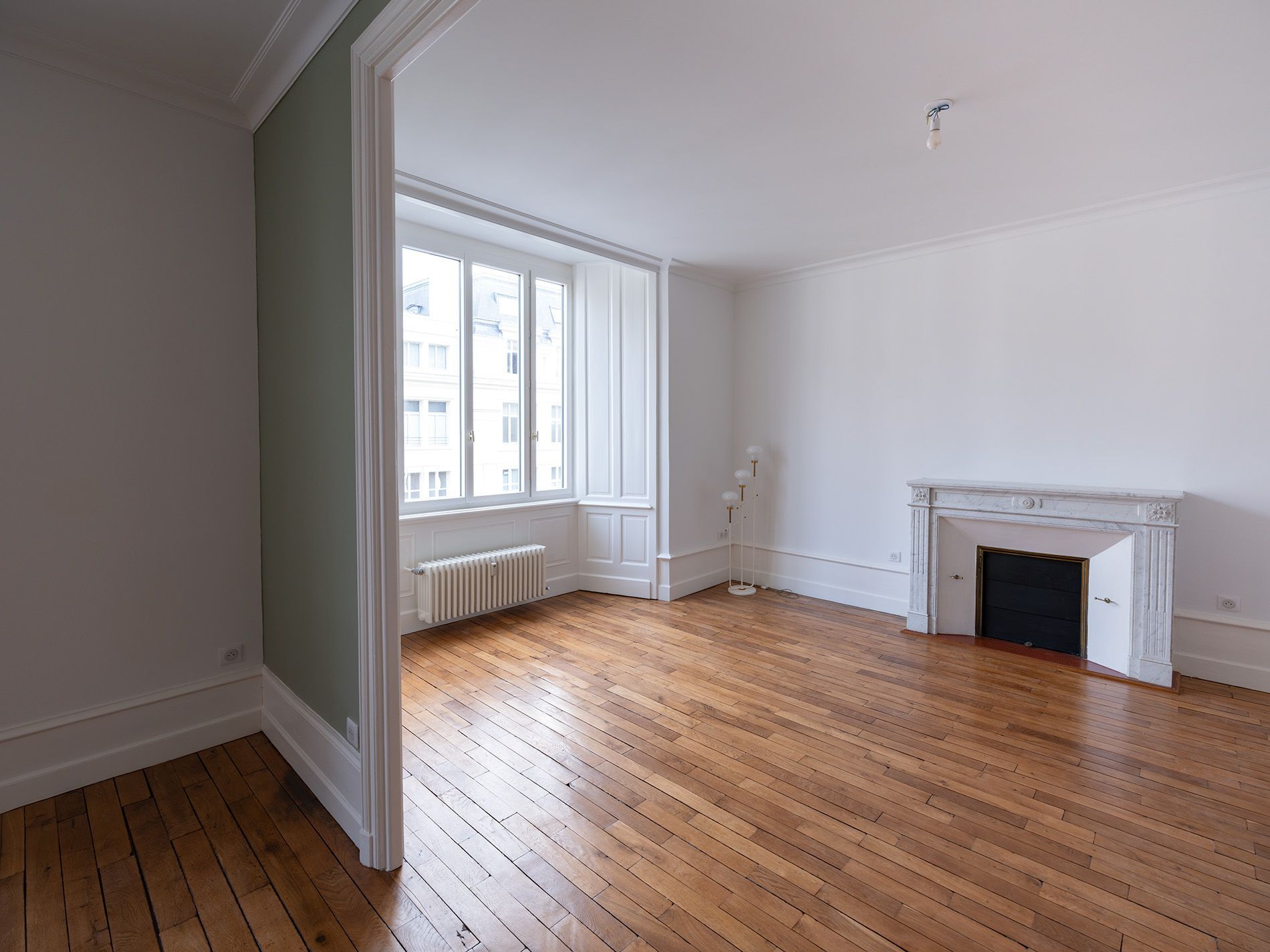 Salon vide avec parquet, grandes fenêtres et cheminée blanche.