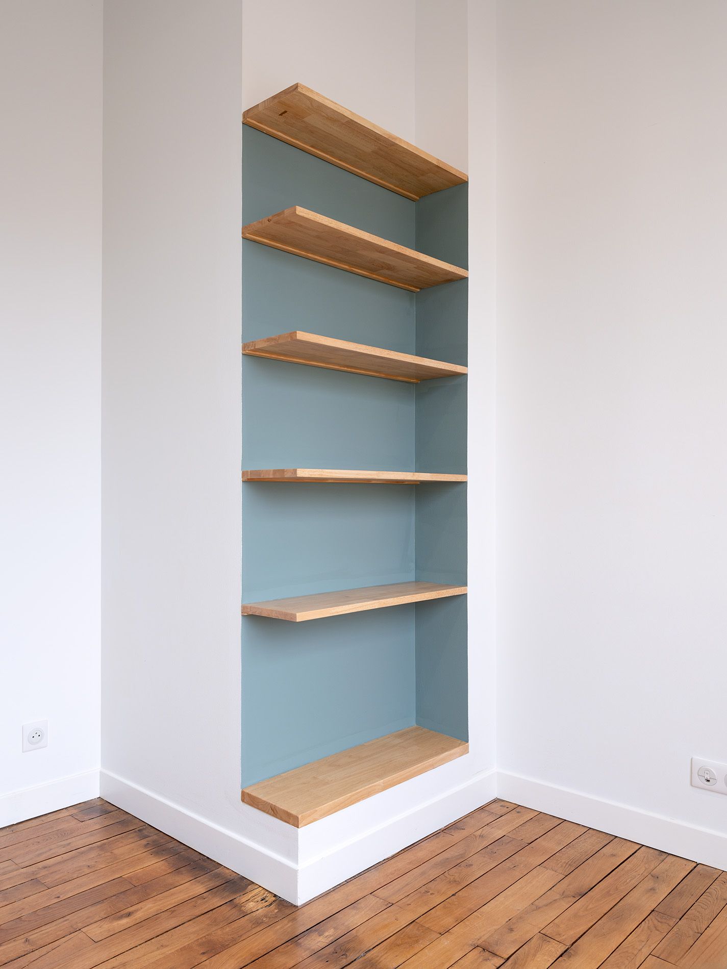 Étagère d'angle intégrée avec des tablettes en bois clair et un panneau arrière bleu dans une pièce blanche.