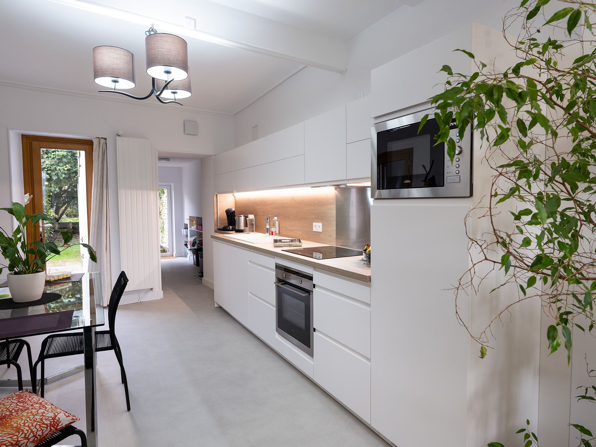 Cuisine moderne blanche en forme de couloir avec coin repas, four encastré et plante d'intérieur près d'une télévision murale