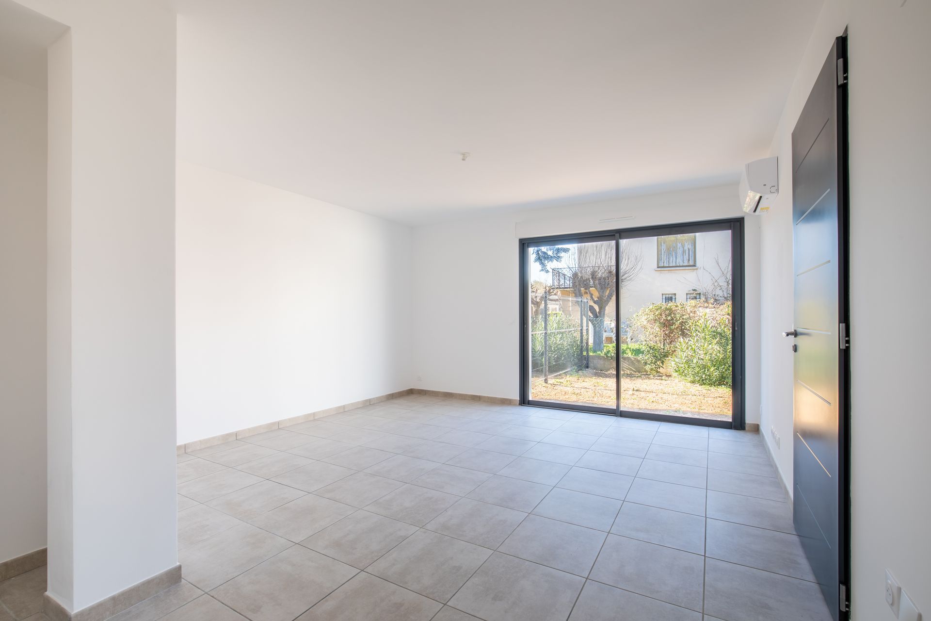 Extérieur de maison moderne avec une porte de garage noire, un enduit gris clair et deux grands arbustes.