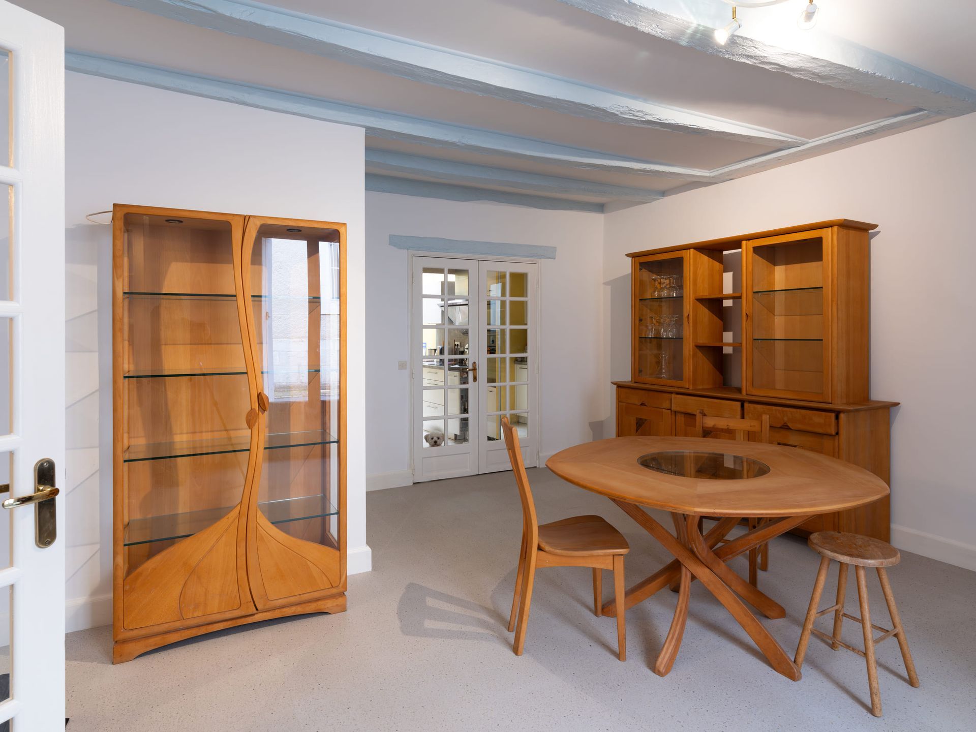 Pièce vide meublée de meubles en bois : étagères, armoire, table ronde, chaises et échelle près d’une porte vitrée.