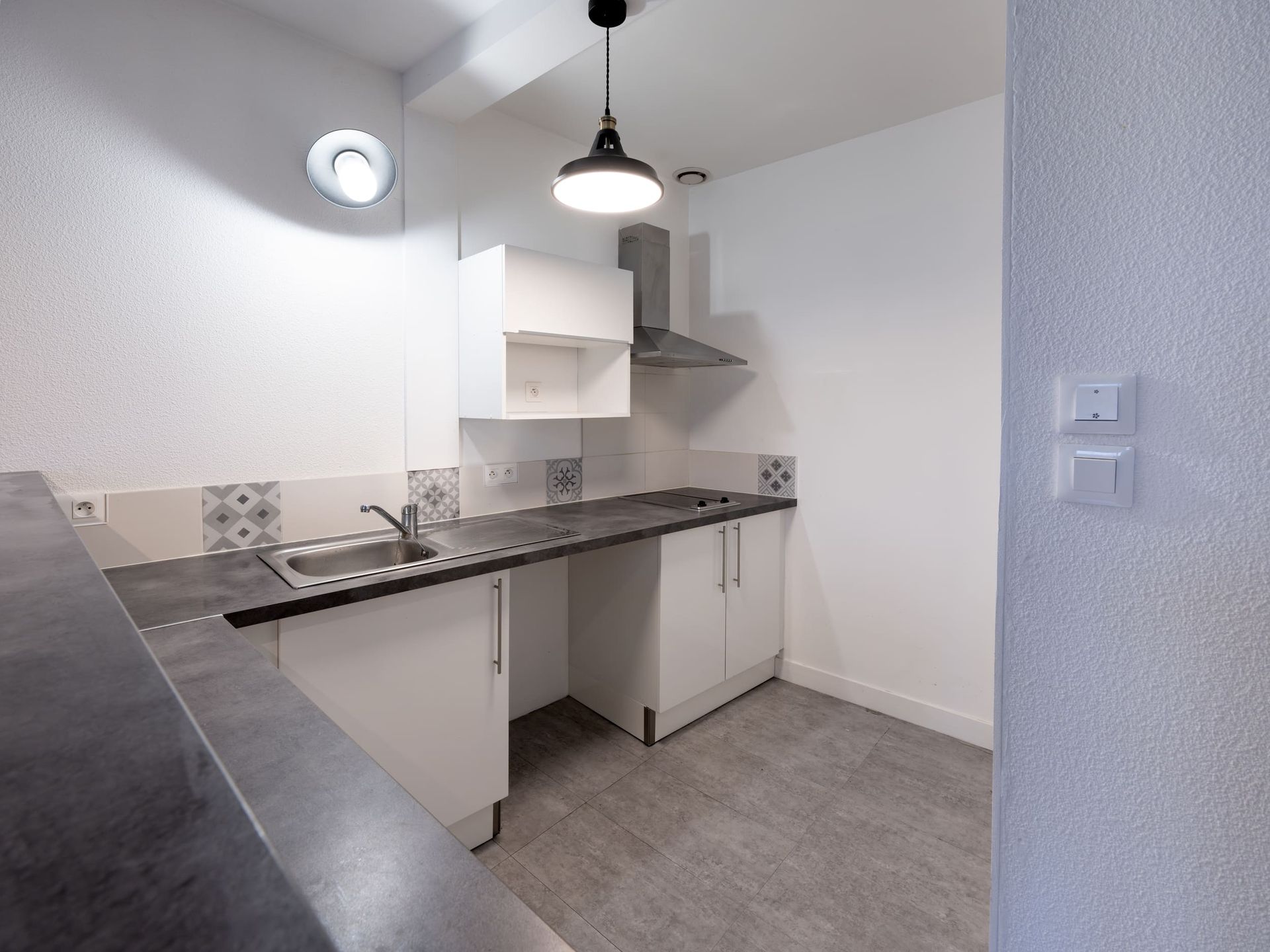 Couloir étroit menant à la cuisine blanche avec comptoirs, armoires, cuisinière, évier et luminaires au plafond gris.