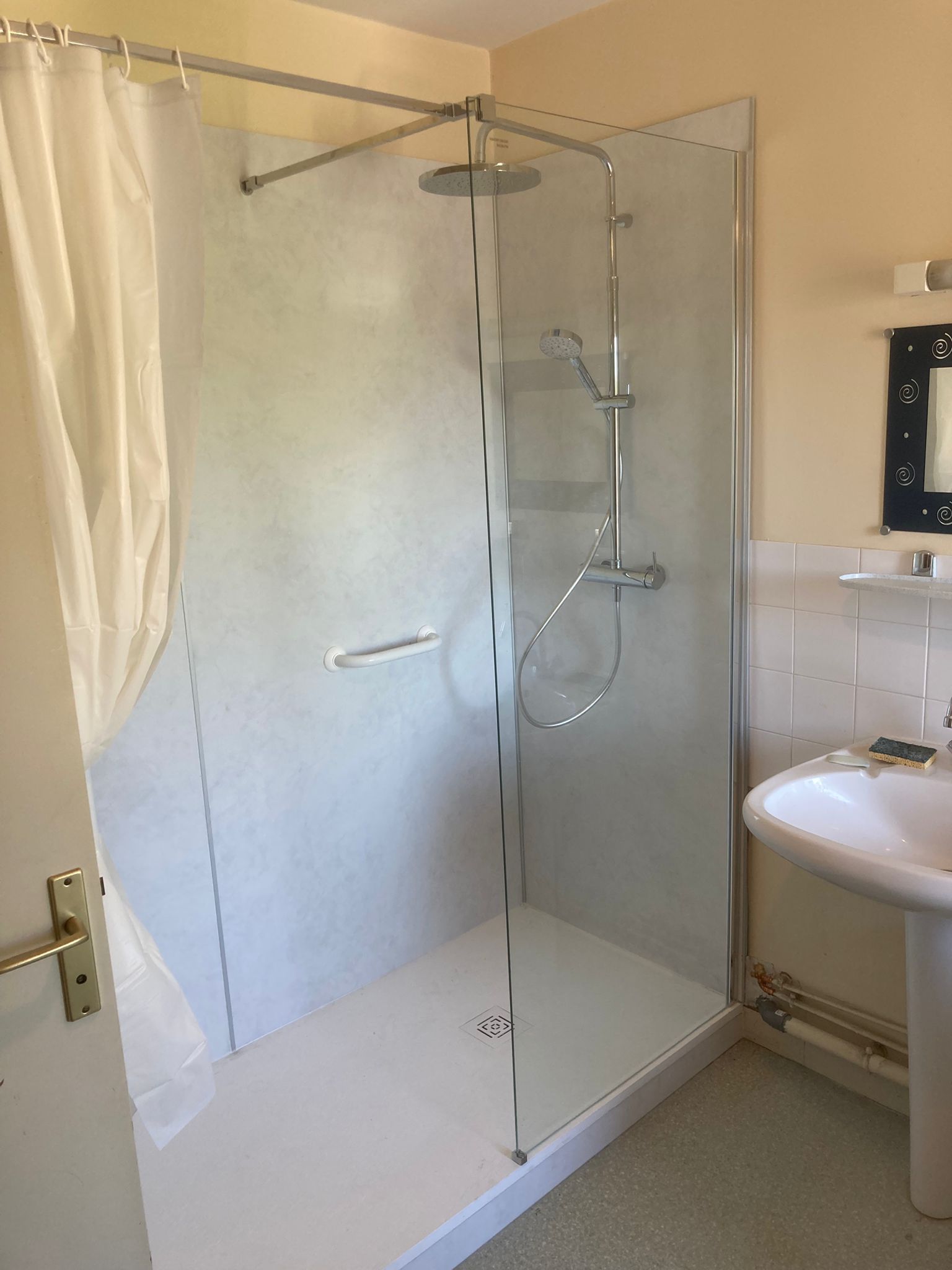 Petite salle de bains avec une cabine de douche en verre, un rideau blanc et un lavabo contre des murs carrelés.