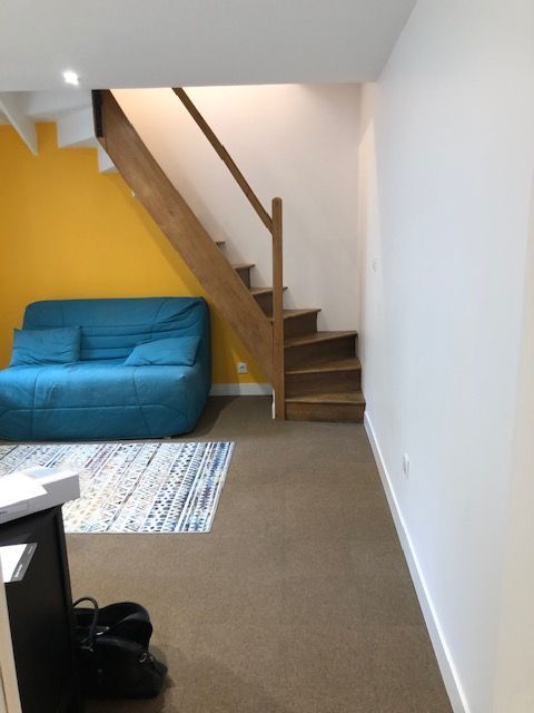 Petit salon avec canapé bleu, escalier, murs blancs et mur d'accent jaune.