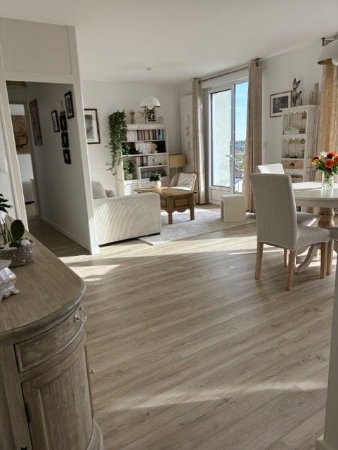 Salon lumineux et ouvert avec parquet clair, meubles blancs, bibliothèque et vue sur balcon.