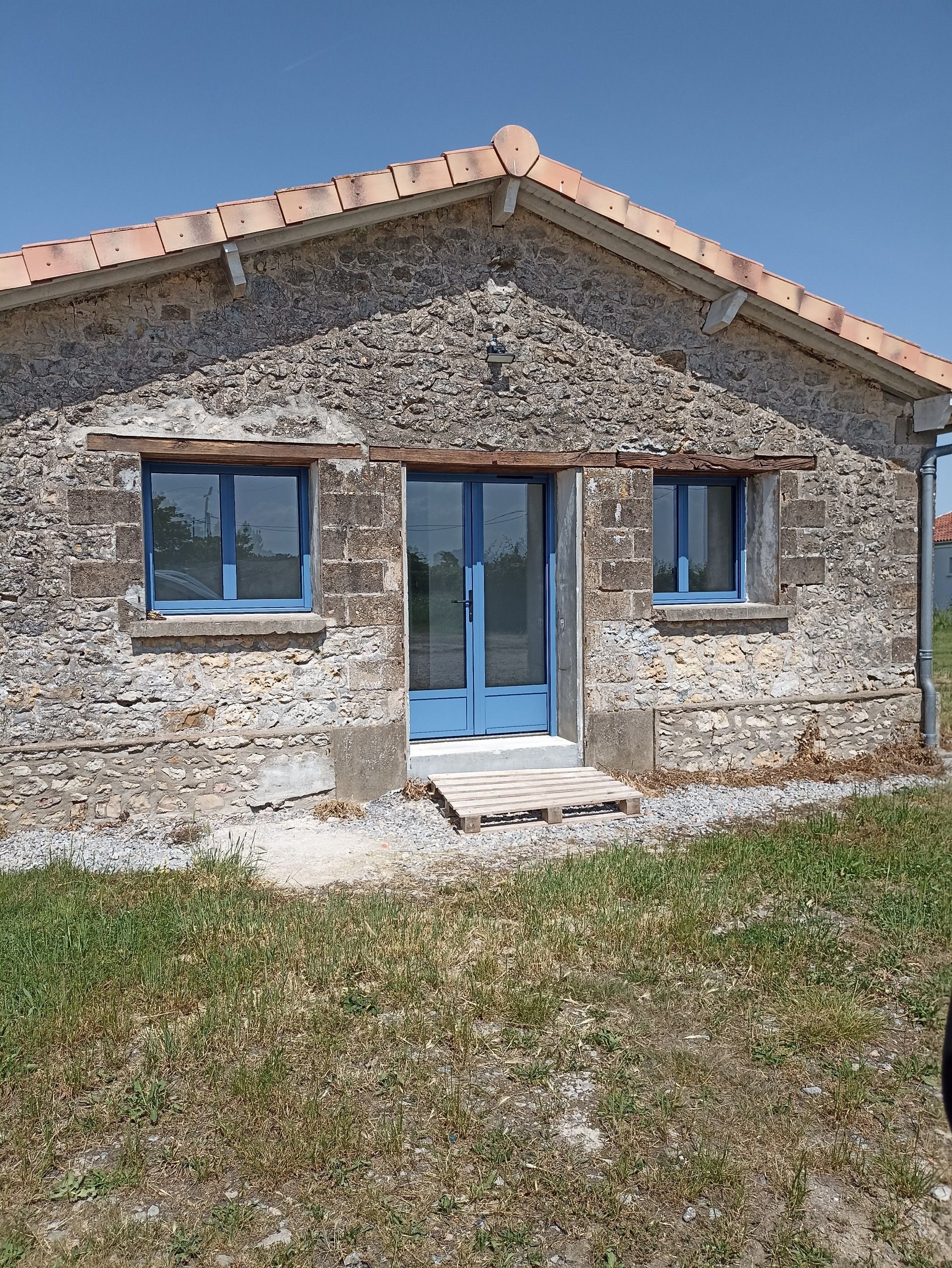 Petite maison rustique en pierre avec une porte d'entrée bleue et deux fenêtres sous un toit de tuiles.