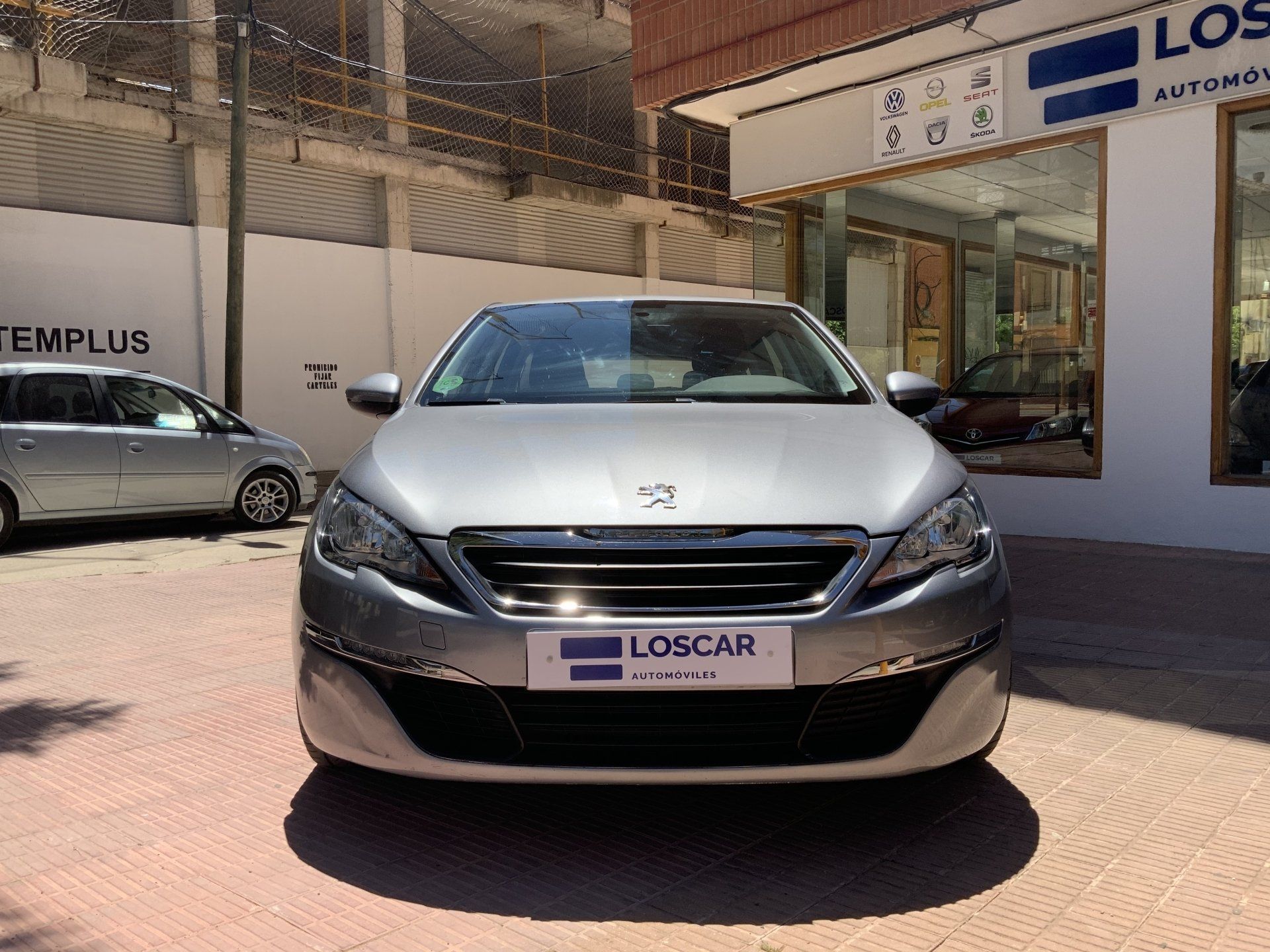 Un Peugeot 308 plateado aparcado frente a un concesionario. El coche está frente a la cámara en un día soleado.