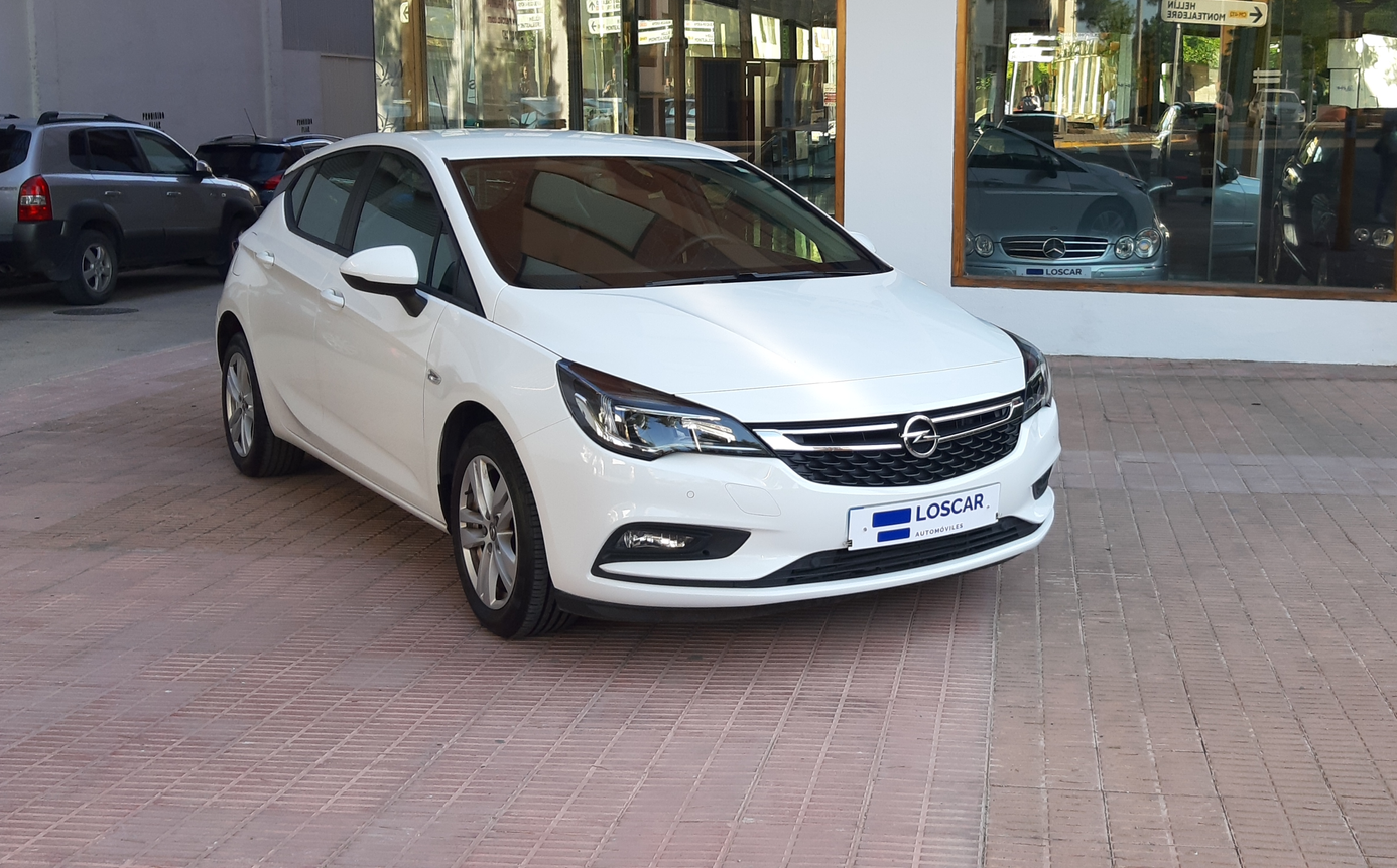 Opel Astra blanco estacionado frente a un edificio con grandes ventanales que reflejan otros coches en el interior.