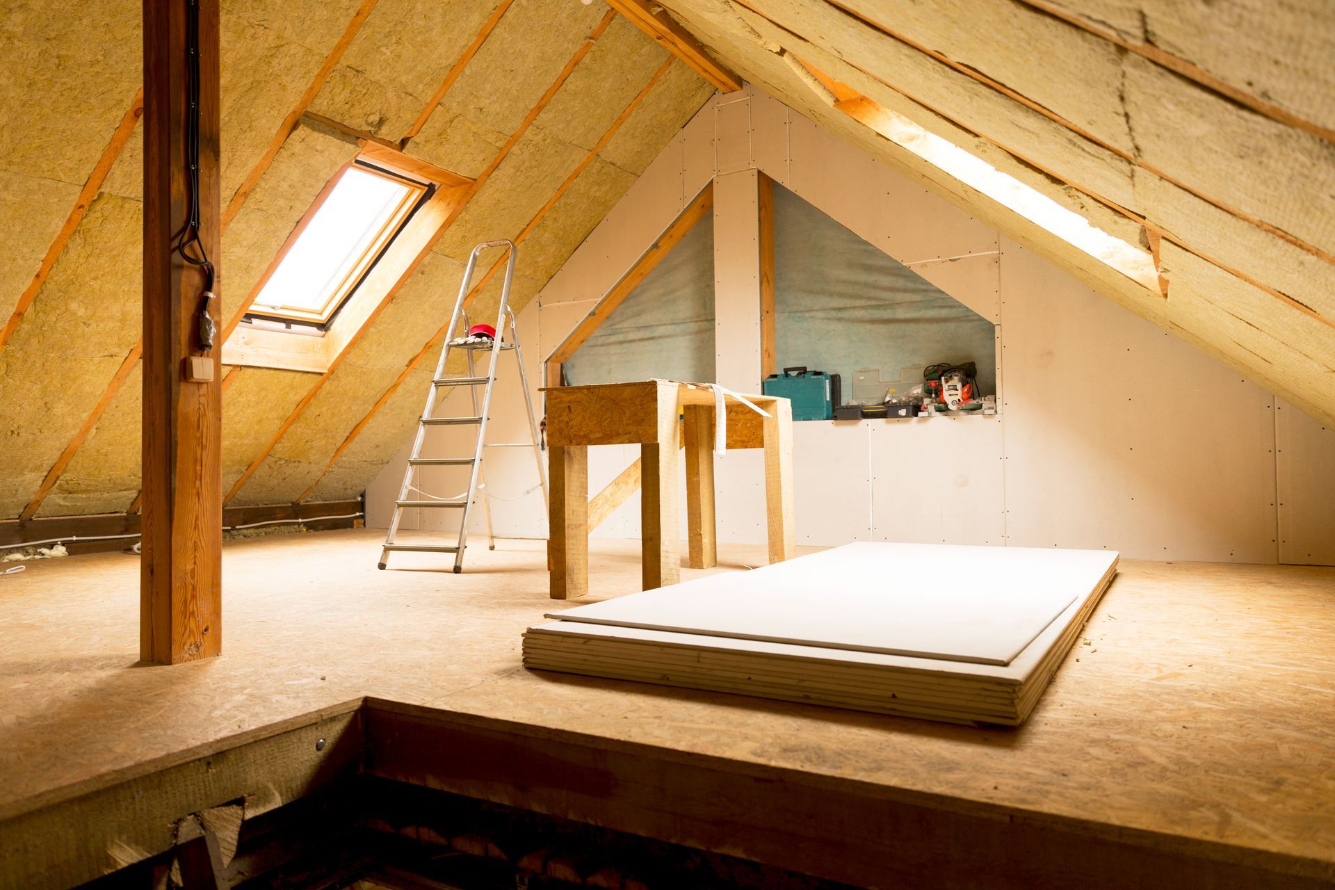 Chambre mansardée partiellement rénovée avec échelle, poutres apparentes et lit surélevé sous un plafond mansardé.