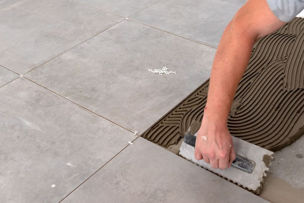 Pose manuelle d'un carreau sur des carreaux de sol gris, à côté d'un lit de colle appliqué à la truelle.