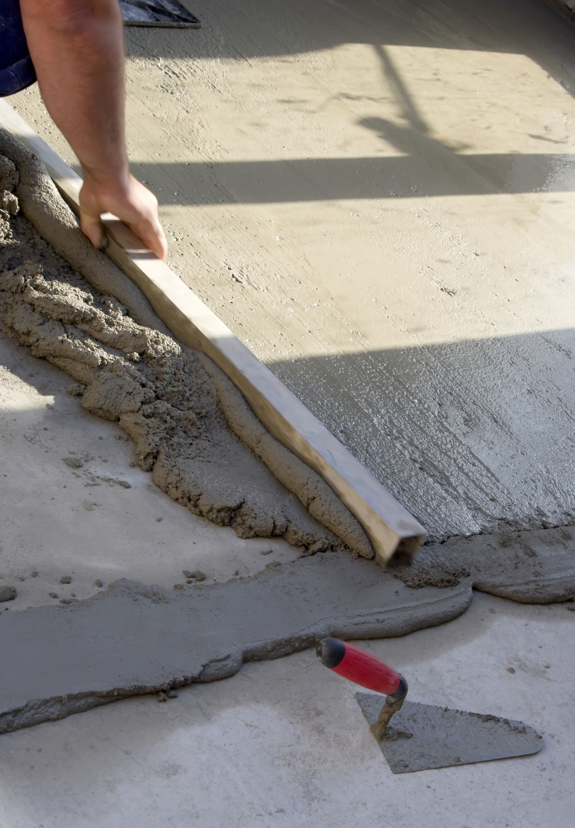 Un ouvrier lisse du béton frais à la truelle, à côté d'une règle sur un chantier de construction.