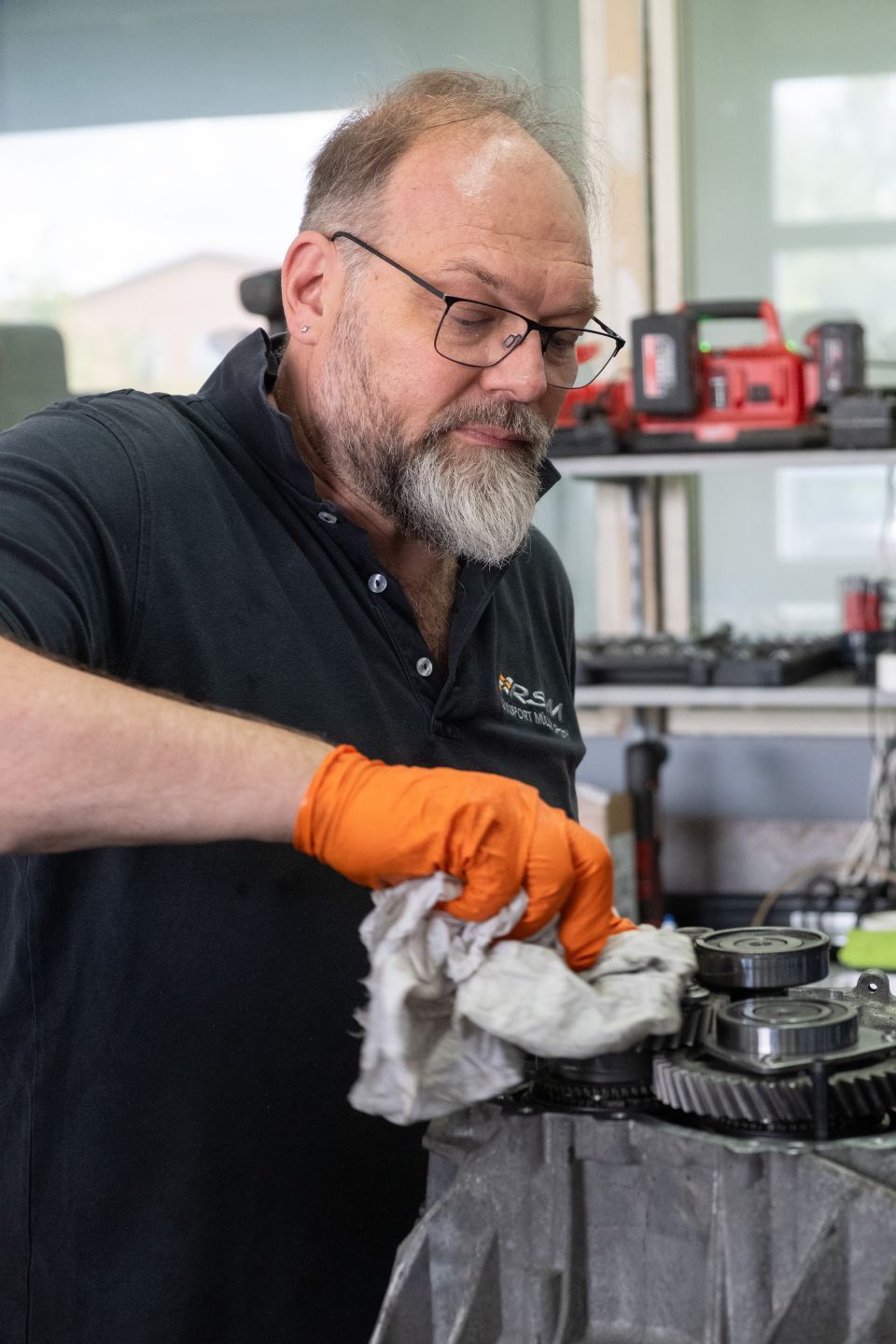 Ein Mechaniker mit orangefarbenen Handschuhen wischt Teile ab und konzentriert sich auf seine Arbeit. Werkstattszene, Werkzeuge im Hintergrund.