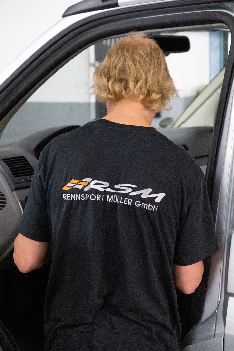 Eine Person mit blonden Haaren und schwarzem T-Shirt steht in einem Auto und arbeitet vermutlich daran.