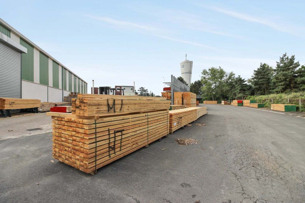 Bois d'œuvre empilé sur des palettes, à l'extérieur d'une scierie. Un bâtiment, des arbres et un château d'eau se dressent à l'arrière-plan.