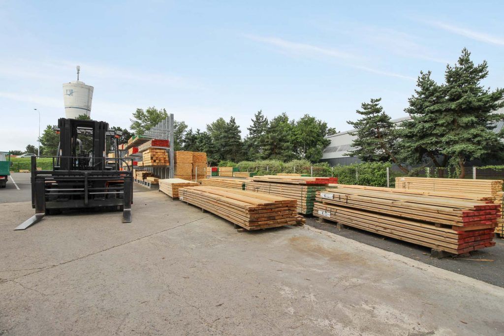 Chariot élévateur chargeant du bois dans une scierie, piles de bois, ciel légèrement couvert.