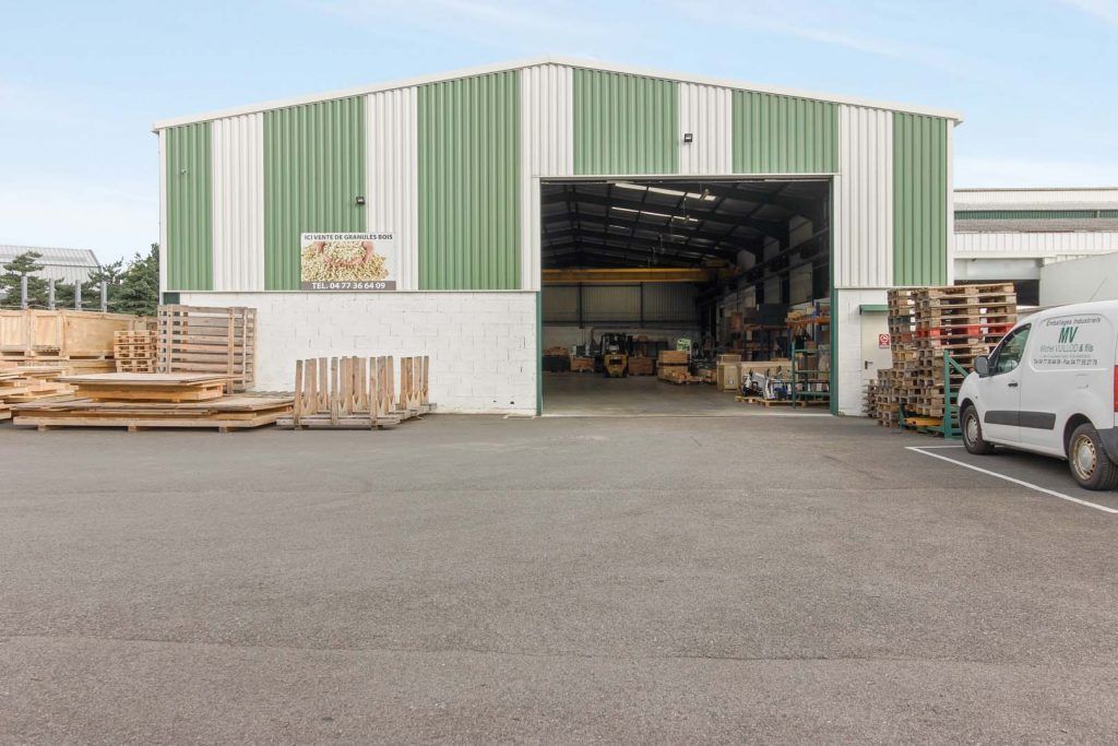 Bâtiment industriel vert et blanc avec porte ouverte, laissant apparaître l'intérieur des entrepôts et des équipements.