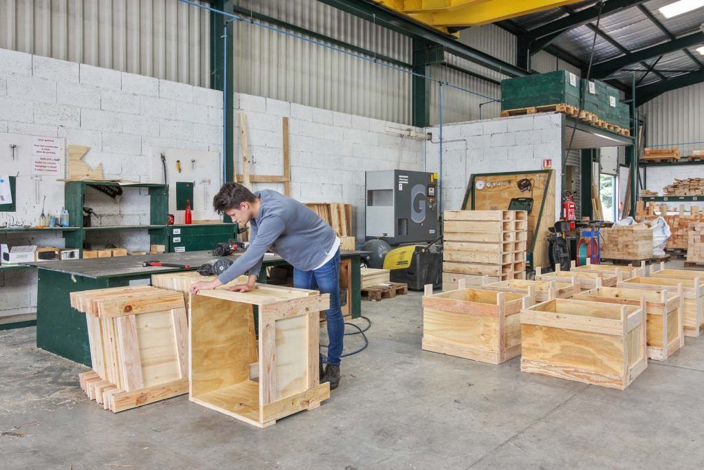 Un homme assemble des caisses en bois dans un entrepôt. Plusieurs caisses terminées sont visibles à proximité.
