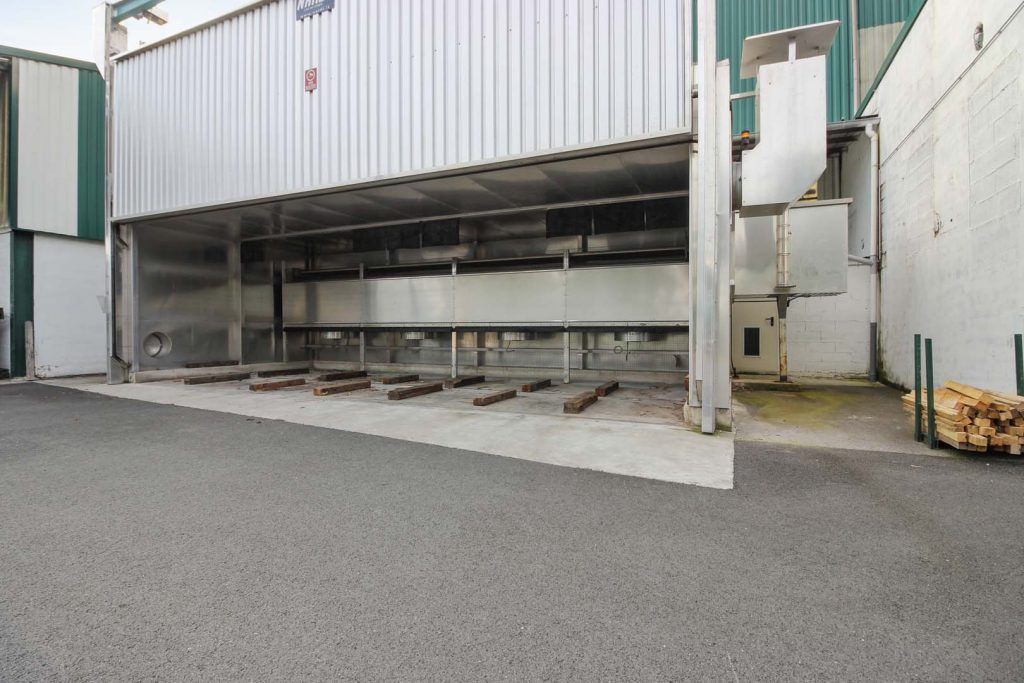 Extérieur d'un grand four industriel à façade ouverte ; bardage métallique, dalle de béton, bûches visibles.