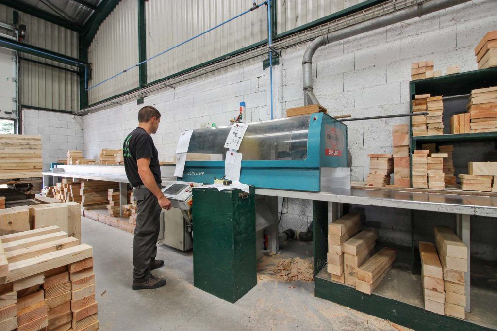 Un homme manipule des machines à bois dans un atelier, entouré de piles de bois.