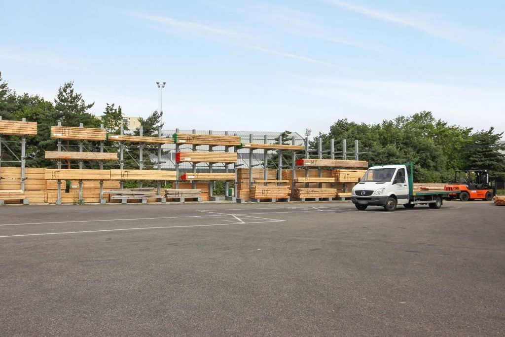 Une scierie avec des étagères en bois, un camion blanc et un chariot élévateur sur un terrain pavé sous un ciel bleu.