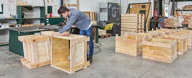 Une personne assemble des caisses en bois dans un atelier, entourée d'autres caisses et d'outils.