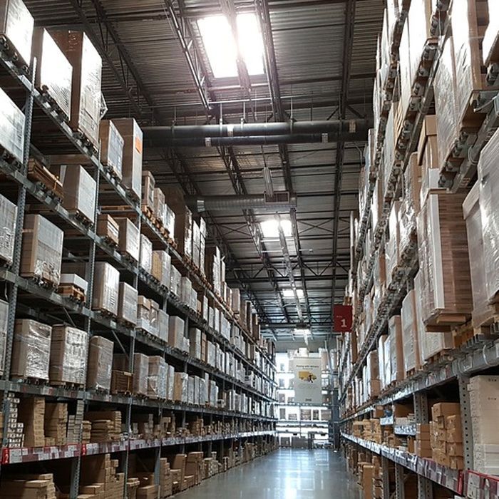 Intérieur d'entrepôt avec de hautes étagères remplies de cartons sous un toit métallique percé de puits de lumière.