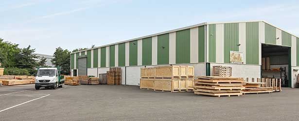 Entrepôt avec bardage à rayures vertes et blanches, une camionnette circulant sur le parking, et des caisses et palettes en bois.