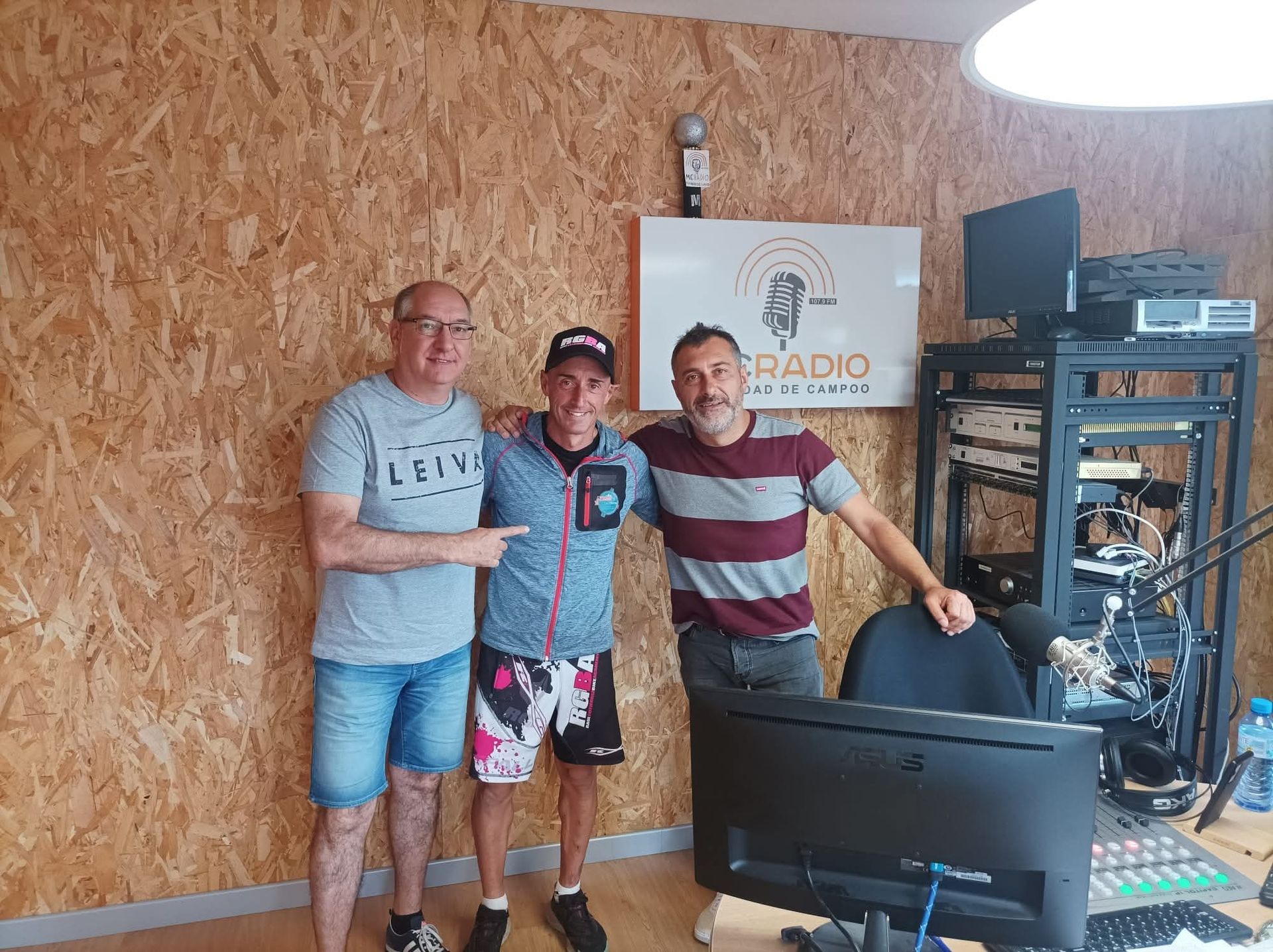 Tres hombres en un estudio de radio. Dos hombres señalan al hombre del centro, todos sonrientes. Se ve el equipo del estudio.