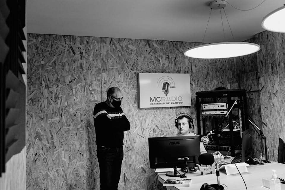 Dos hombres en un estudio de radio: uno hablando por un micrófono, el otro de pie con los brazos cruzados.