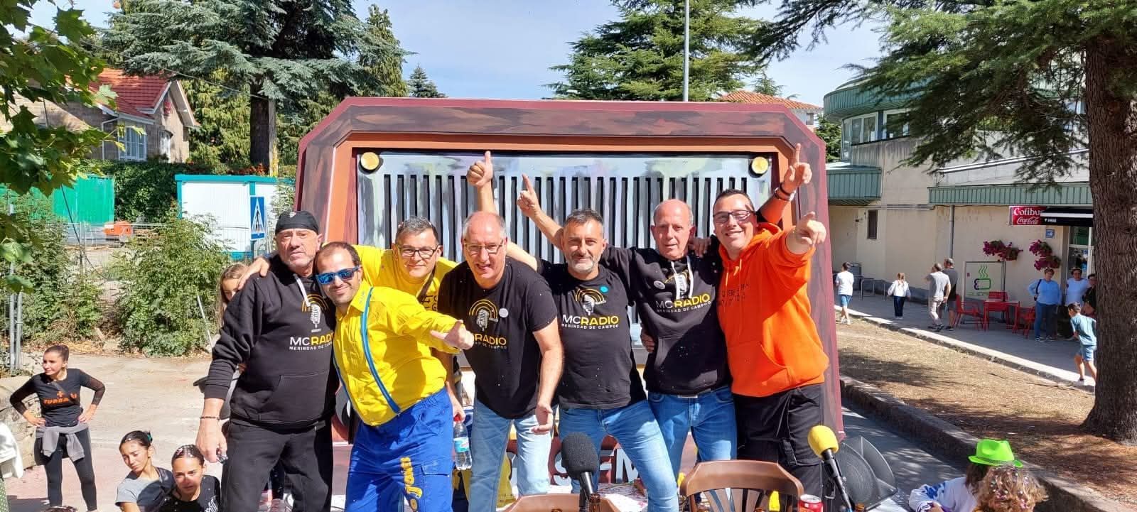 Un grupo de hombres posa frente a un gran soporte de radio, sonriendo y celebrando al aire libre.