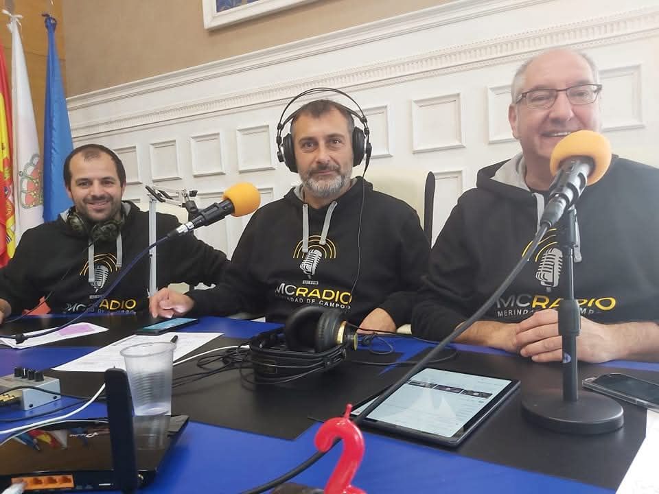 Tres hombres en un estudio de radio con auriculares, hablando por micrófonos. Sudaderas negras y mesa azul.