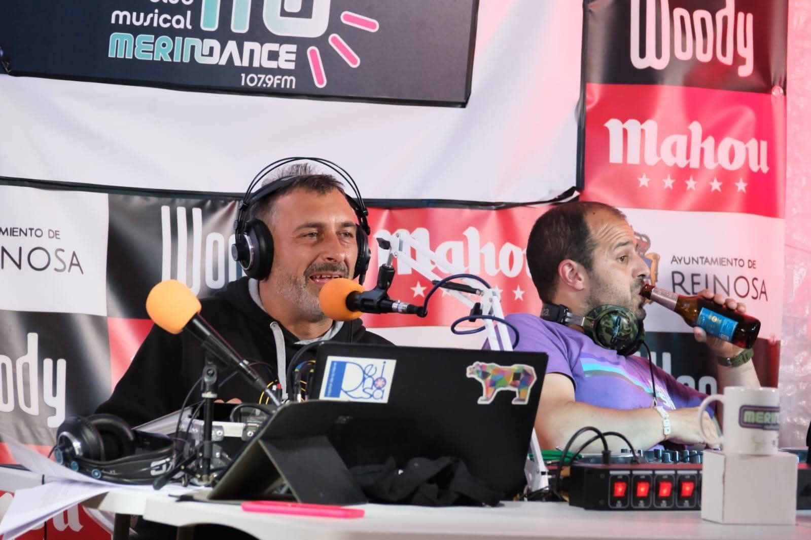 Dos hombres en una radio; uno habla por un micrófono, el otro bebe cerveza, ambos sonriendo.