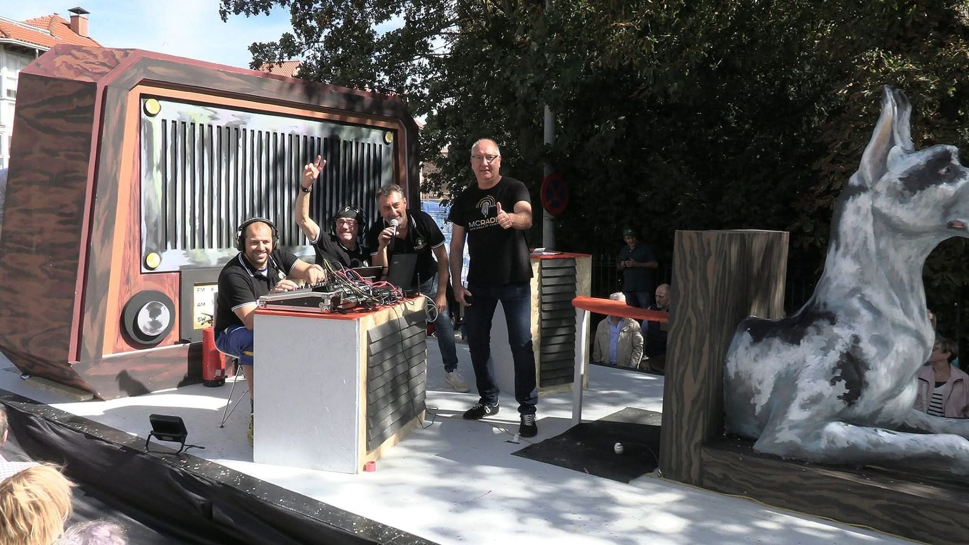 Un grupo de hombres en una carroza de desfile con una estatua de un perro y una fachada de camión, sonriendo y gesticulando al aire libre.