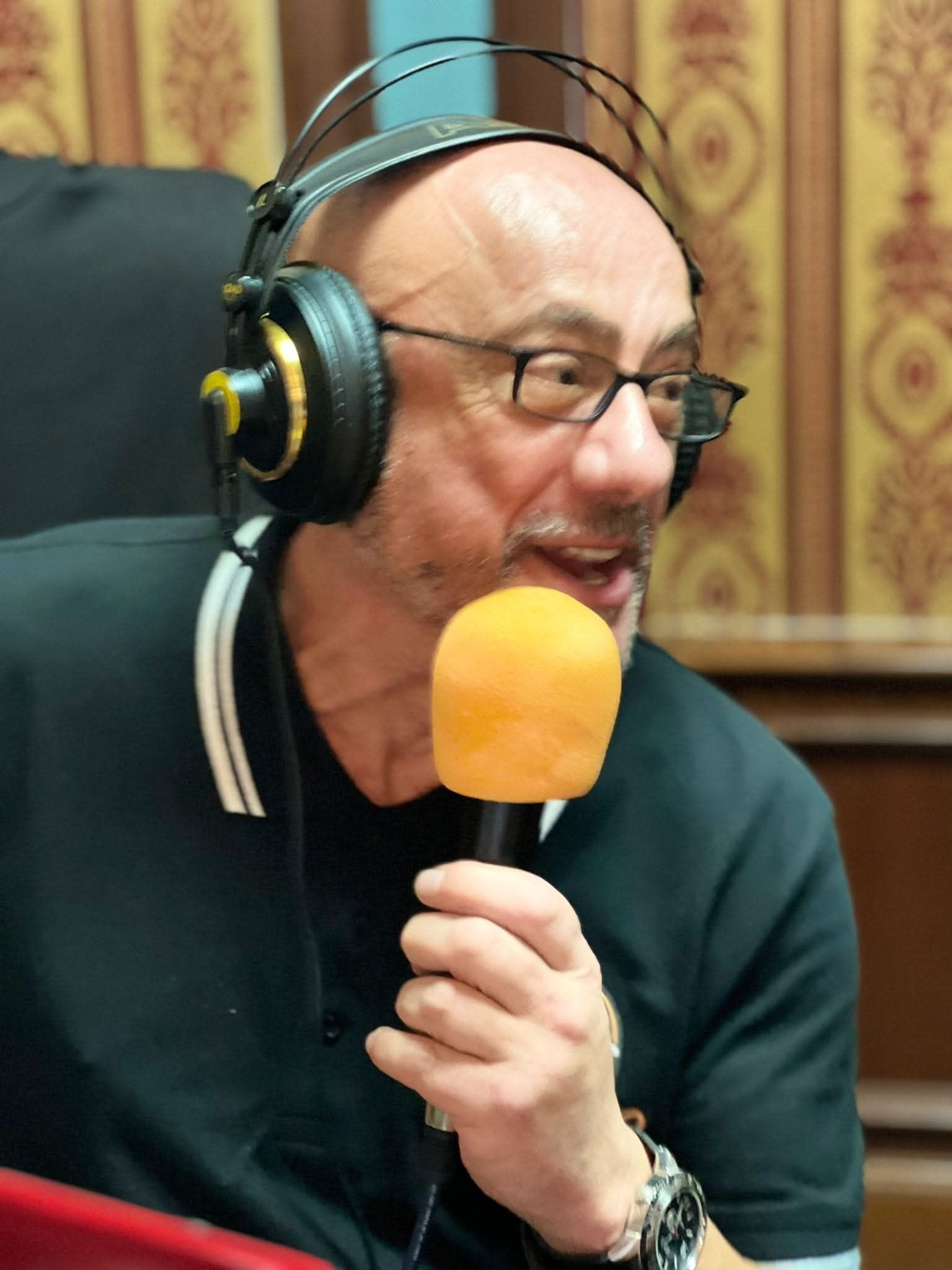 Hombre con gafas y auriculares, sosteniendo un micrófono, hablando en una habitación con paredes estampadas.