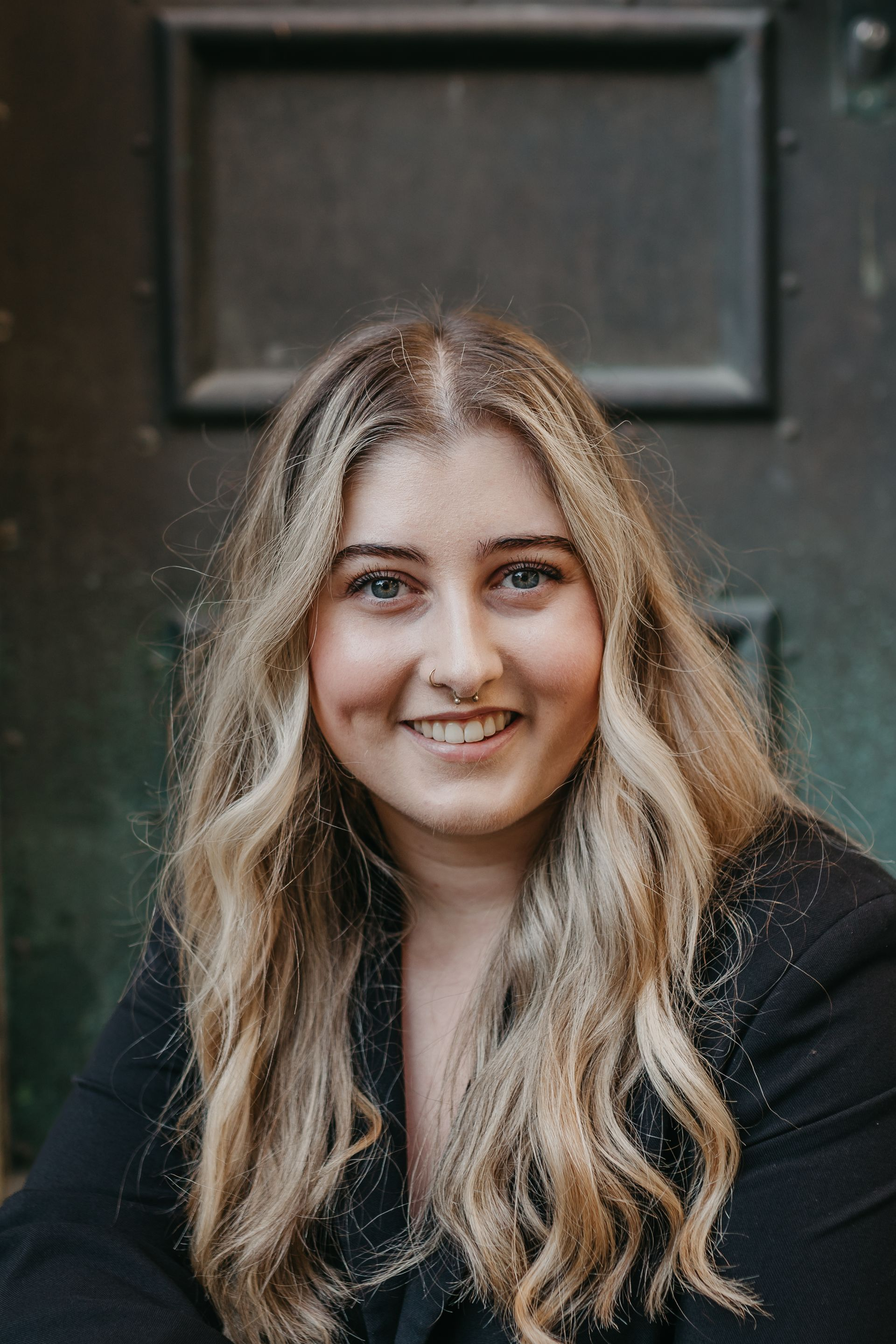 Junge Frau mit welligem braunem Haar lächelt im Freien, trägt ein weisses Hemd, im Hintergrund ist Herbstlaub zu sehen.