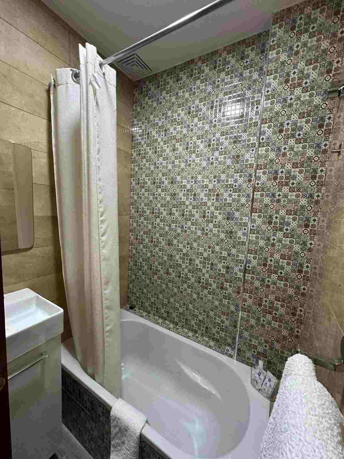 Baño pequeño con cortina de ducha, azulejos decorativos en la pared, bañera, lavabo y portarrollos de papel higiénico.