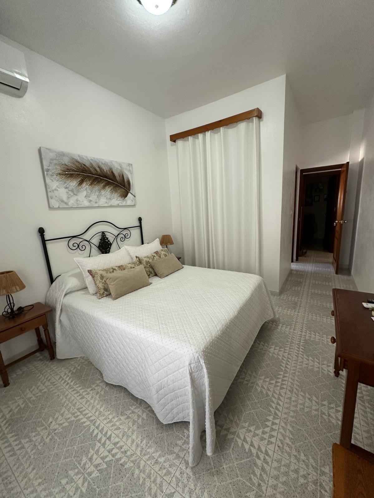 Habitación luminosa con ropa de cama blanca, detalles en color beige, un escritorio de madera y una puerta al fondo.