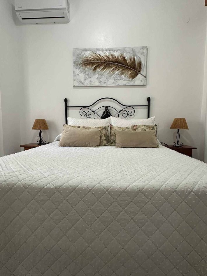 Dormitorio con una colcha blanca acolchada, almohadas beige, lámparas de noche y un cuadro en la pared sobre el cabecero.