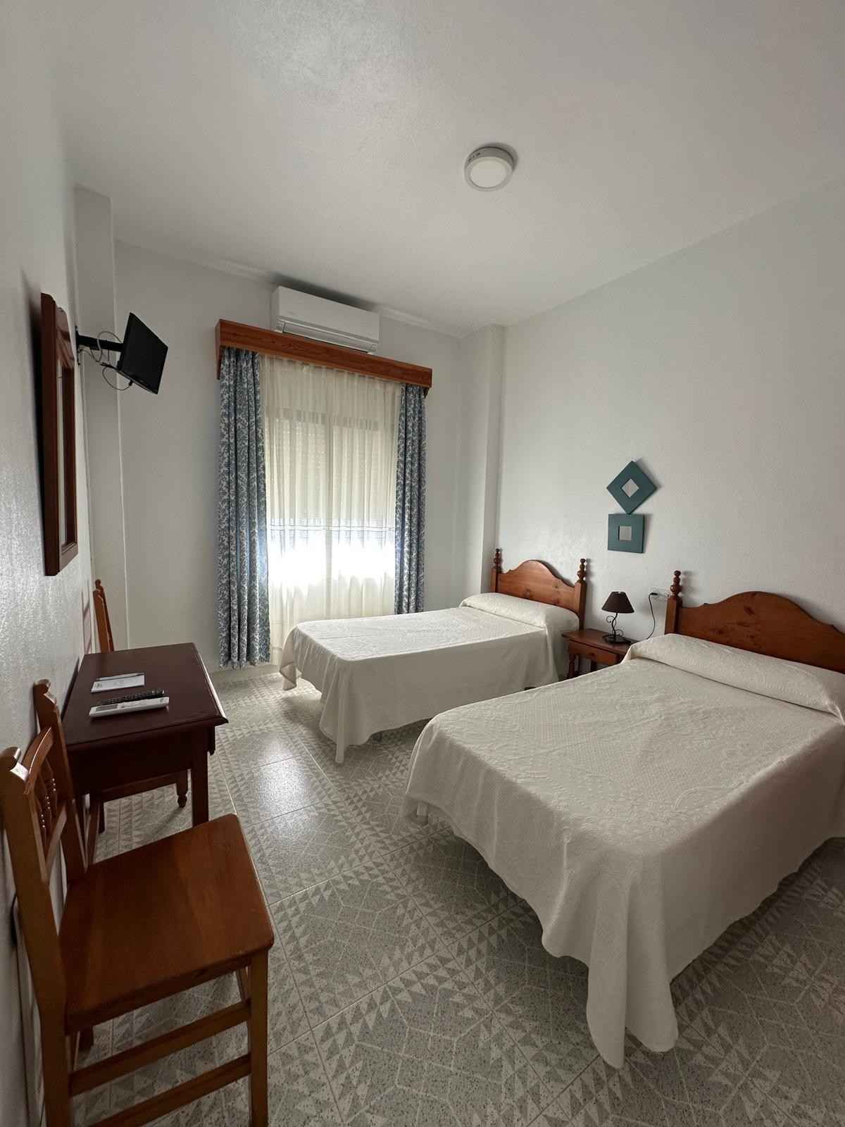 Habitación pequeña con dos camas individuales, ventana, muebles de madera y colchas blancas.