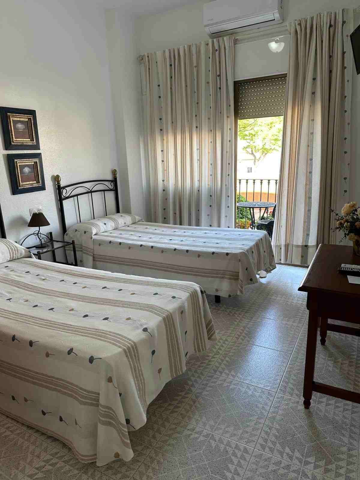 Habitación pequeña con dos camas individuales, colchas estampadas y una puerta que da al balcón con cortinas transparentes.