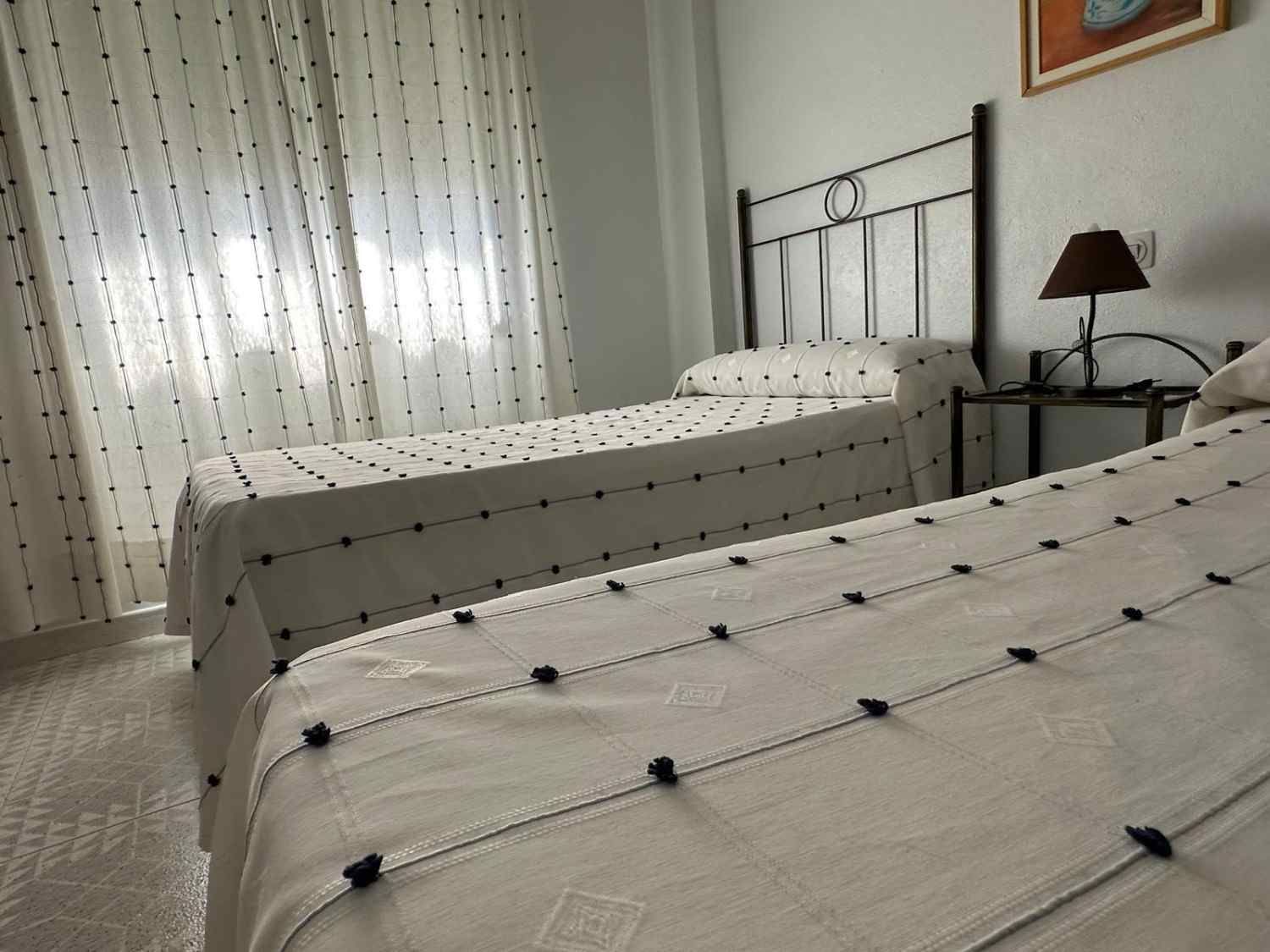 Dos camas individuales en una habitación luminosa con cortinas transparentes, ropa de cama blanca y una lámpara de noche.