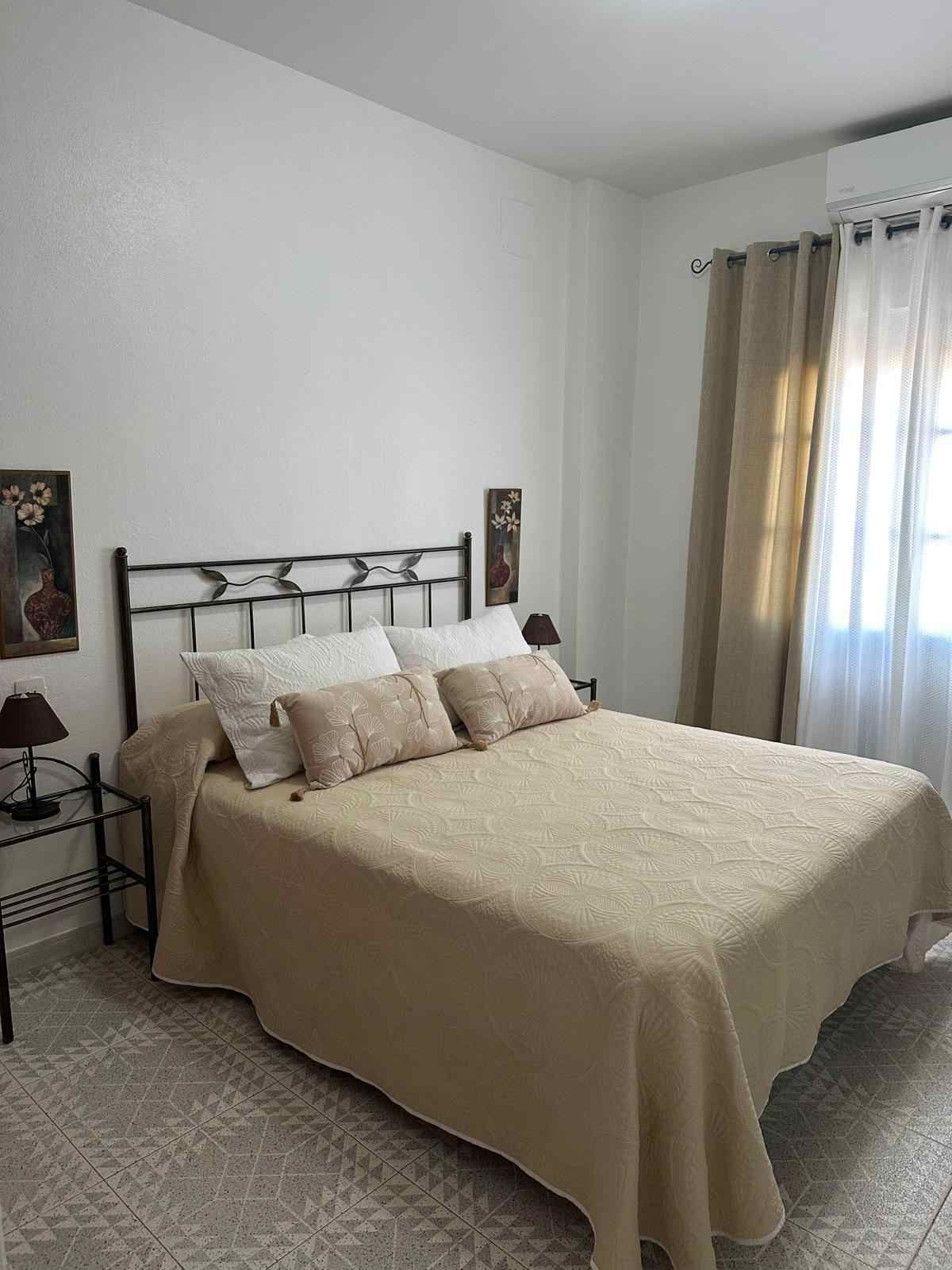 Habitación luminosa con una colcha beige, paredes blancas y una ventana con cortinas.