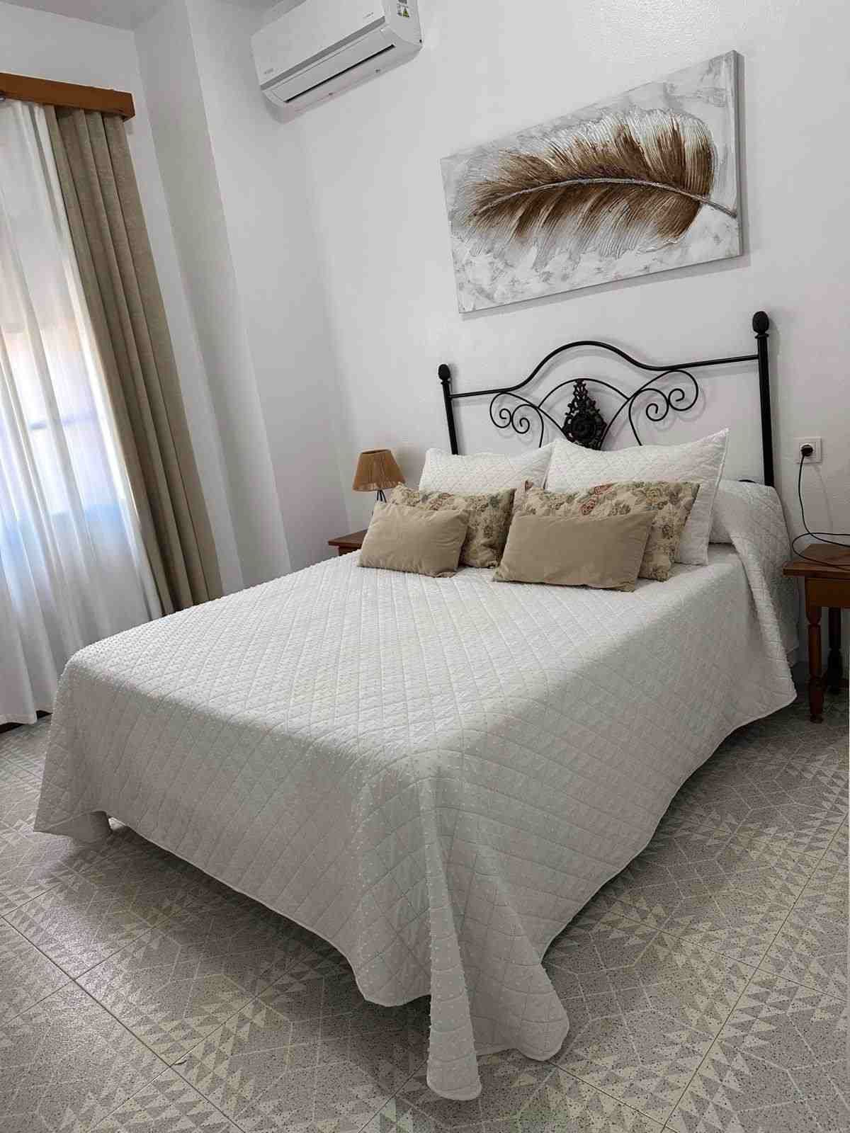 Habitación luminosa con cama blanca, almohadas beige, alfombra estampada, cuadros en la pared y cortinas en la ventana.