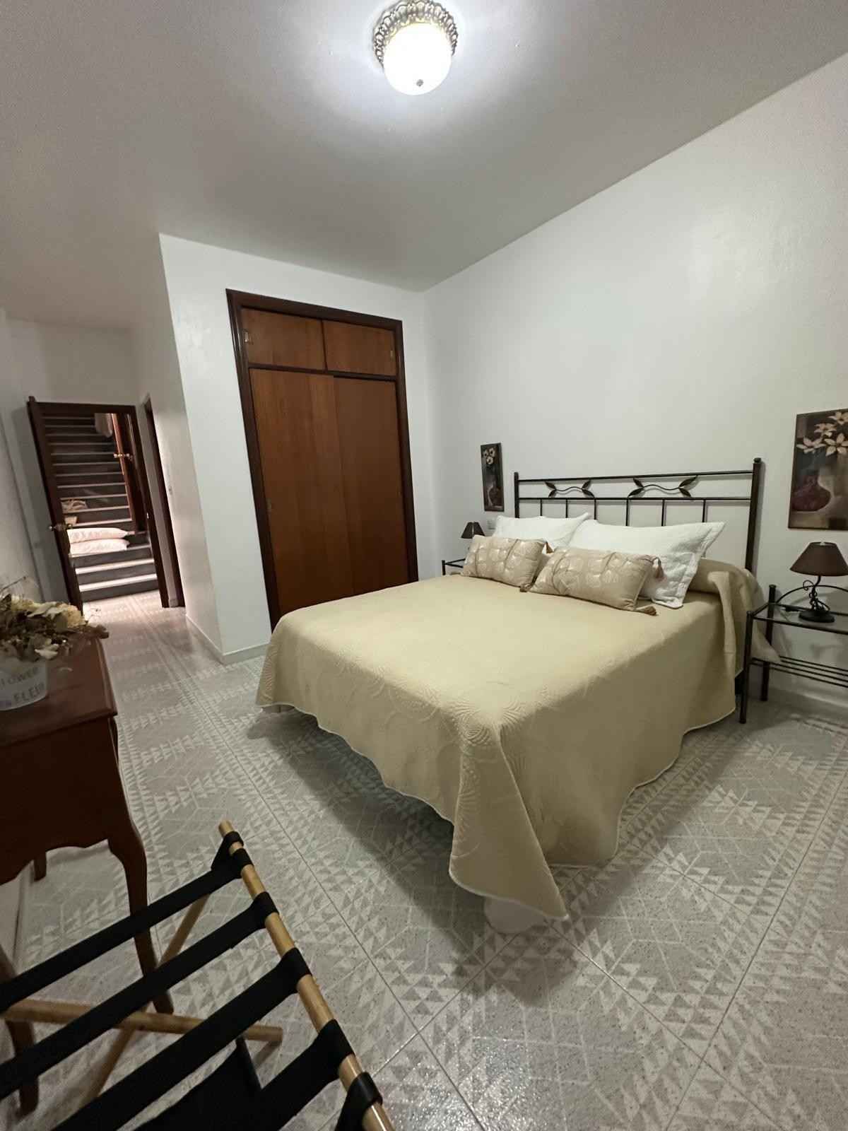 Habitación pequeña con cama de metal, ropa de cama beige, armario de madera y baldosas estampadas en el suelo.