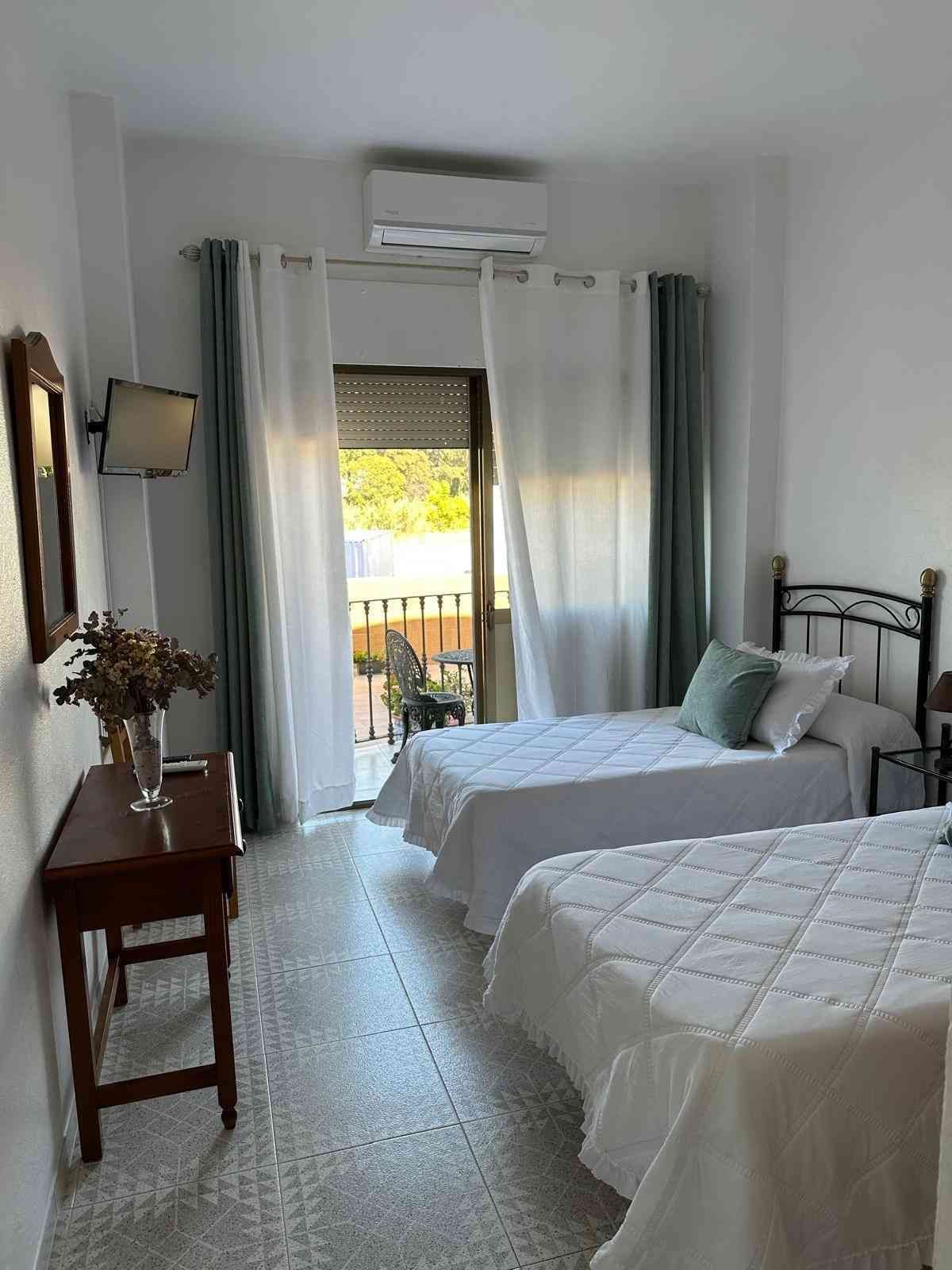 Habitación luminosa con dos camas, puerta de acceso al balcón y escritorio de madera junto a la pared.