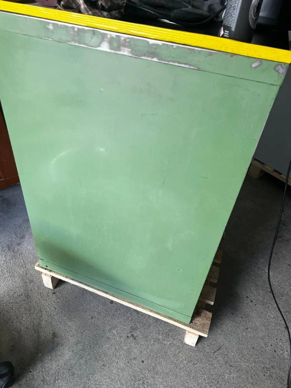 Ein salbeigrüner Metallschrank mit leuchtend gelber oberer Zierleiste steht auf einer kleinen Holzpalette auf einem Betonboden.