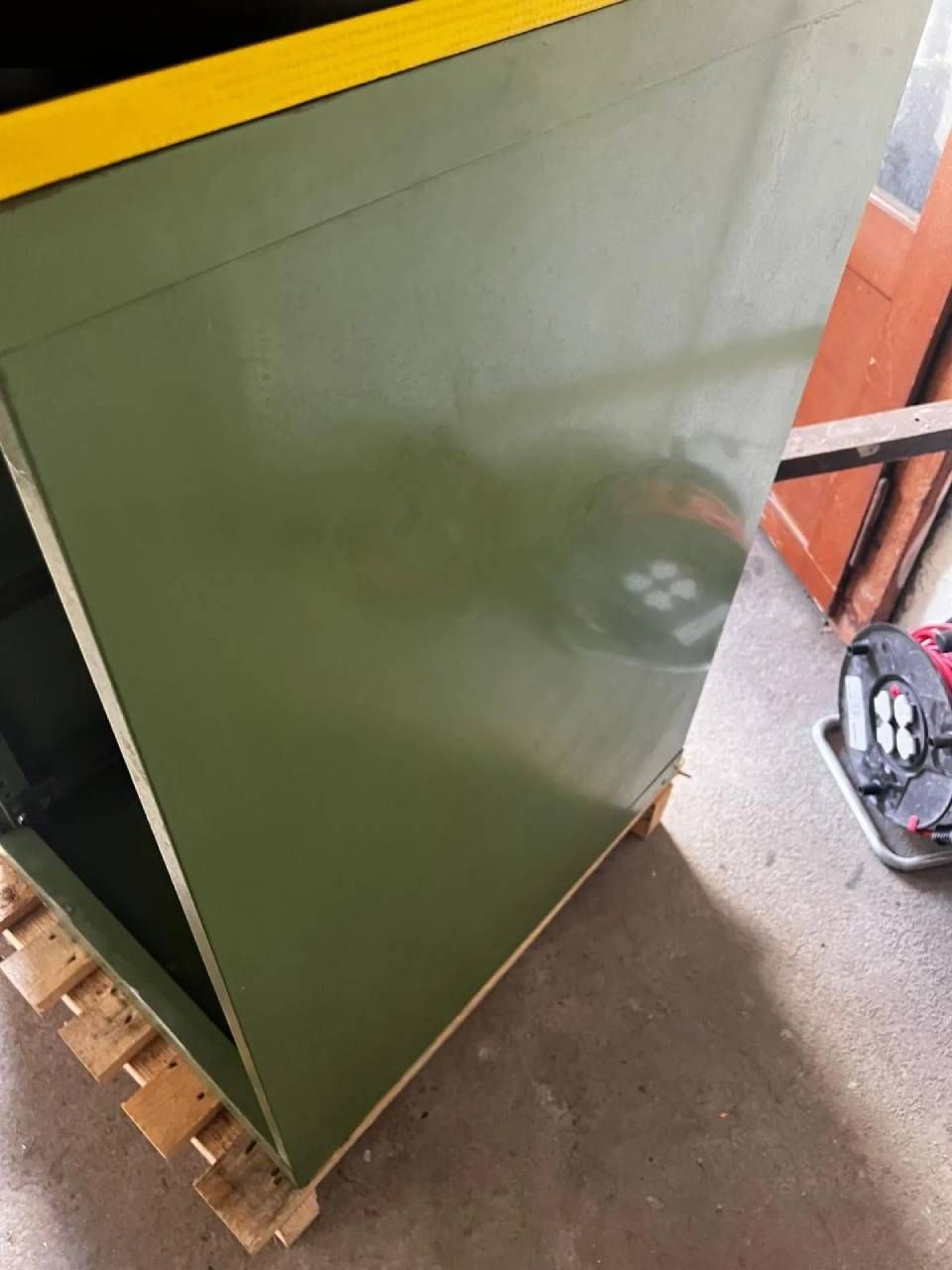 In einer Werkstatt steht auf einer Holzpalette ein olivgrüner Metallschrank mit einer markanten gelben Oberkante.