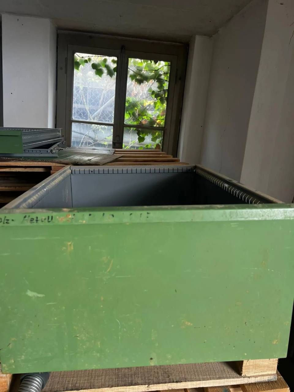 Ein grüner, rechteckiger Bienenstock mit gekerbten Kanten, der auf einer Holzpalette vor einem Fenster steht.